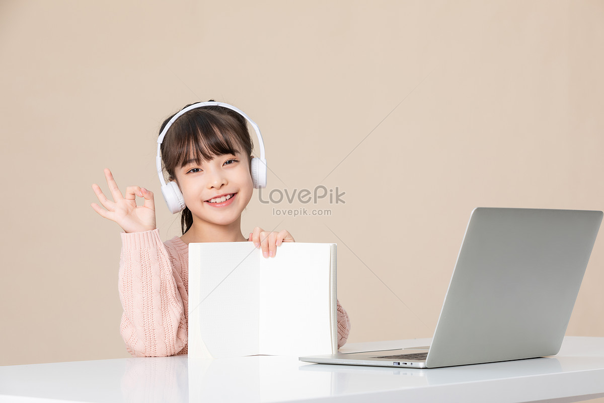 Little girl gestures OK to the camera holding a homework book, computer desk, and homework, book HD Photo